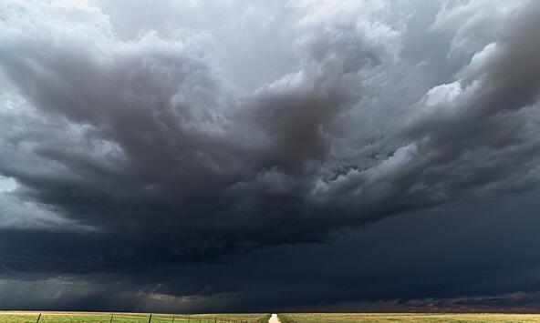 Storm clouds over a long straight road ahead, flanked by fields 